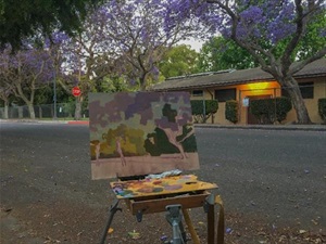 Painting in progress on easel with paint box set up in street with blooming jacaranda trees in background, painting in progress by Alexey Steele to be titled "Jacaranda Bloom. Culver City" 2021, oil on linen panel, 16in-x-20in