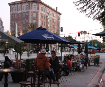 Photograph showing people seated at outdoor dining tables with umbrellas in closed street area along Culver Blvd with Culver Hotel in background