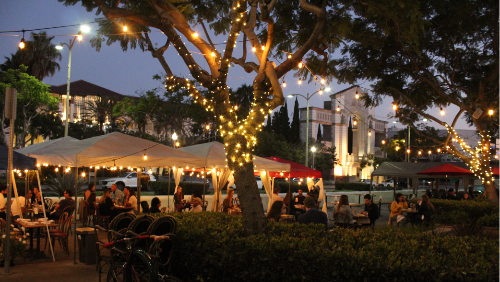 Nighttime photograph showing lighted trees and people seated at outdoor dining tables under lighted tents located in closed street area along Culver Blvd with City Hall in background