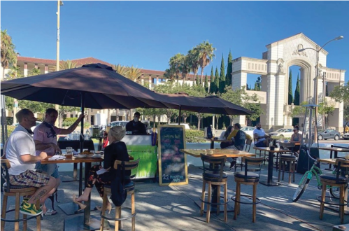Photograph showing people seated at outdoor dining tables and umbrellas located in closed street area along Culver Blvd with City Hall in background