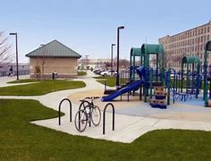 Photograph of bicycle secured to u-shaped bike rack at a park, next to playground with large play structure and buildings in the background