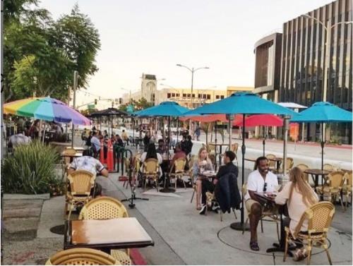 Photograph showing people seated at outdoor dining tables and umbrellas located in closed street area along Culver Blvd with retail buildings in background