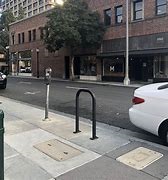 Photograph of bicycle secured to u-shaped bike rack along a city street, next to parked car and parking meter with buildings in background