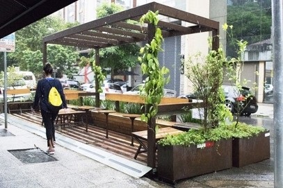 Photograph of outdoor dining installation in city street along street curb, showing tables, benches and plants in planter boxes, covered by trellis, with woman with yellow backpack walking by.
