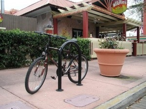 Photograph of bicycle secured to u-shaped bike rack, along street, in front of public building, next to large planter
